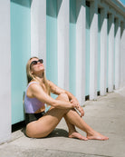 Eine Frau mit Sonnenbrille und dem Nova Badeanzug von Main Design sitzt auf einem sonnigen Bürgersteig, lehnt sich an eine pastellblaue und weiße Wand und genießt lächelnd mit geschlossenen Augen das warme Wetter.