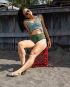 Eine Frau mit Sonnenbrille und grünem Main Design Nova Badeanzug aus recycelten Materialien sitzt mit ausgestreckten Beinen und sandigen Füßen auf einer roten Kiste am Strand und lehnt sich mit zur Seite geneigtem Kopf zurück.