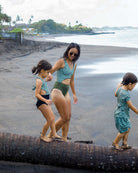 Eine Frau im Main Design Nova Badeanzug aus recycelten Materialien hilft einem jungen Mädchen, auf einem Palmenstamm am Strand zu balancieren, während ein Junge vorausläuft. Sie genießen den Blick aufs Meer und die sich wiegenden Palmen im Hintergrund.