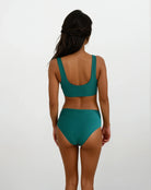 Eine Frau mit langem, dunklem Haar steht mit abgewandtem Gesicht da und trägt die blaugrüne Alice Bikinihose von Main Design aus nachhaltigen Materialien mit einem Oberteil mit U-Ausschnitt und einer hoch taillierten Hose vor einem schlichten weißen Hintergrund.