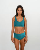 Eine Frau mit langen dunklen Haaren steht vor einem weißen Hintergrund. Sie trägt einen türkisfarbenen Sport-BH und die Alice Bikinihose von Main Design aus nachhaltigen Materialien. Sie blickt mit neutralem Gesichtsausdruck direkt in die Kamera.