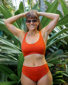 Eine lächelnde Frau in einem Main Design Charm Surf Bikini Oberteil und Sonnenbrille steht mit den Händen auf dem Kopf inmitten üppiger grüner tropischer Pflanzen.