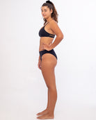 Eine Frau steht barfuß vor einem weißen Hintergrund in einem nachhaltigen Bliss-Bikinihosen-Set von Main Design. Sie blickt nach rechts, hat die Hände an den Seiten und trägt eine hoch taillierte Hose. Ihr Haar ist zu einem Pferdeschwanz zurückgebunden und verkörpert sowohl Stil als auch umweltfreundliche Prinzipien.