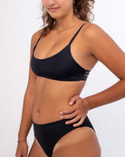 Eine Frau mit langen, lockigen Haaren trägt eine schwarze Bliss-Bikinihose von Main Design. Sie blickt mit einem leichten Lächeln in die Kamera, während sie vor einem schlichten weißen Hintergrund steht.