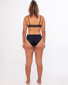 Eine Person mit lockigem Haar steht mit dem Rücken zur Kamera und trägt einen schwarzen Riemchen-Sport-BH und die hoch taillierte Bliss Bikinihose von Main Design. Der Hintergrund ist schlicht weiß.