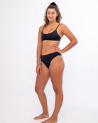 Eine Person mit lockigem Haar lächelt, während sie in der nachhaltigen Bliss Bikinihose von Main Design posiert, mit hoher Taille für Stil und Komfort, vor einem schlichten weißen Hintergrund. Sie steht in entspannter Haltung mit einer Hand auf der Hüfte.