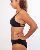 Eine Frau im Profil steht vor einem schlichten Hintergrund und trägt die Bliss Bikinihose von Main Design. Ihre Haare sind mit einem Haarband zurückgebunden. Sie stemmt die Hände in die Hüften und blickt nach vorne.