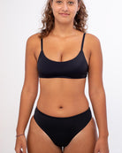 Eine Frau mit lockigem Haar steht lächelnd vor einem schlichten weißen Hintergrund und präsentiert die nachhaltige Bliss-Bikinihose mit hohem Bund von Main Design.