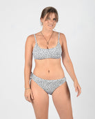 Eine Frau steht selbstbewusst vor einem schlichten Hintergrund und trägt einen weißen Bikini mit schwarzem Punktmuster. Sie hat ihre Haare zurückgebunden, eine Halskette um den Hals und lächelt sanft in die Kamera. Dabei präsentiert sie die Blush Surf Bikinihose von Main Design aus umweltfreundlichem Stoff.