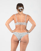 Eine Frau mit heller Haut steht mit dem Rücken zur Kamera. Sie trägt einen nachhaltigen Bikini in Weiß mit schwarzen Punkten, ergänzt durch die Blush Surf Bikinihose von Main Design. Ihr Haar ist mit einem passenden Haargummi zurückgebunden und gibt den Blick auf eine Halskette frei. Der Hintergrund ist schlicht und hell.