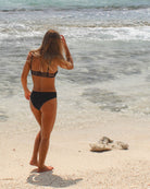 Eine Person mit langen Haaren und einem schwarzen Bikini mit Ember Bikini Hose von Main Design geht an einem Sandstrand, an dem verstreut kleine Steine liegen, zum Meer. Das Wasser ist klar und sanfte Wellen nähern sich dem Ufer.