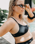 Eine Frau in einem Sassy Bikini-Top von Main Design, das aus recycelten Materialien hergestellt wurde und einen sportlichen Schnitt aufweist, sitzt an einem Pool und rückt ihre Sonnenbrille zurecht. Sie hat lange blonde Haare, eine Halskette und trägt ein schwarzes Haargummi am Handgelenk.