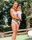 Eine Frau in einer Flair-Bikinihose von Main Design steht an einem Pool und hält ein kleines Kind in einem lila- und cremefarbenen Badeanzug im Arm. Beide lächeln, mit Bäumen und einem Zaun als ruhigem Hintergrund.
