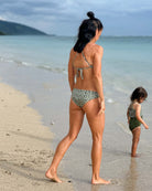 Eine Frau in einer gemusterten Luxe Bikini Hose von Main Design spaziert am Sandstrand entlang, während ein Kind in einem passenden Badeanzug am Wasser steht, mit dem Meer und den fernen Hügeln im Hintergrund.