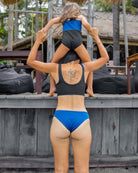 Eine Frau trägt die Main Design Luxe Bikini Hose in Blau und Schwarz, steht mit dem Rücken zur Kamera und trägt ein junges Mädchen in einem passenden Badeanzug auf den Schultern in der Nähe von Holzstufen und Palmen.