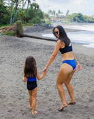 Eine Frau und ein junges Mädchen in passenden Luxe Bikini-Hosen von Main Design halten sich an einem Sandstrand an den Händen, mit Palmen und Meereswellen im Hintergrund, während die Frau lächelnd zurückblickt.