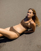 Eine Frau im Sassy Glitter Bikini Oberteil von Main Design liegt auf der Seite im dunklen Sand, das Sonnenlicht hebt ihre Haut und ihr Haar hervor, während die ruhige Strandszene den Schimmer ihrer stilvollen Bademode unterstreicht.