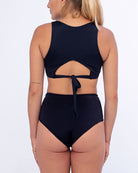 Eine Frau mit blondem Haar und Pferdeschwanz steht mit dem Rücken zur Kamera und zeigt Main Designs Posh Bikini Hose, einen schwarzen zweiteiligen Badeanzug aus recycelten Materialien. Das Oberteil zum Binden am Rücken und die hoch taillierte Hose unterstreichen ihren Stil, während ein kleines Tattoo ihren linken Arm vor dem schlichten weißen Hintergrund ziert.