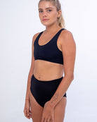 Eine Person mit langen, zu einem Pferdeschwanz gestylten Haaren trägt ein schwarzes ärmelloses Oberteil und dazu eine hoch taillierte **Posh Bikinihose** von **Main Design**, die aus recycelten Materialien gefertigt ist. Sie steht vor einem schlichten Hintergrund, zeigt Tattoos auf ihren Armen und behält einen ruhigen Gesichtsausdruck bei.