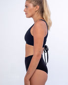 Eine Frau mit langen blonden Haaren zu einem Pferdeschwanz, einem roten Haargummi, einem eleganten schwarzen Sportoberteil und der Posh Bikinihose von Main Design aus recycelten Materialien steht im Profil vor einem schlichten weißen Hintergrund. Ihre Arme sind mit kleinen Tattoos verziert.