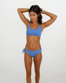 Eine Frau trägt das Charm Surf Bikini-Oberteil von Main Design in Blau und posiert vor einem weißen Hintergrund mit den Händen im Haar und betont UPF 50+ Sonnenschutz für optimalen Schutz beim Wassersport.