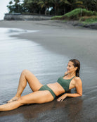 Eine Frau, die die Main Design Serene Surf Bikini Hose trägt, die aus recycelten Materialien hergestellt und in grüner Farbe chlorresistent ist, entspannt sich auf nassem Sand am Meer mit Bäumen im Hintergrund.