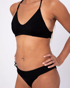 Eine Person modelliert die Main Design Serene Surf Bikini Hose, posiert mit einer Hand auf der Hüfte vor einem schlichten weißen Hintergrund und hebt einen gepiercten Bauchnabel und einen Ring am Finger hervor.