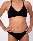 Eine Frau modelliert die Main Design Serene Surf Bikini Hose in Schwarz, die aus recycelten Materialien hergestellt ist, und steht mit den Händen in den Hüften vor einem schlichten weißen Hintergrund. Nur ihr Körper von den Schultern bis zu den Oberschenkeln ist zu sehen.