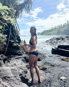 Eine Frau, die die Main Design Tide Surf Bikini Hose mit verstellbarem Kordelzug trägt, steht auf vulkanischen Felsen am Meer und hält Flossen. Üppiges Grün und Bäume sind im Hintergrund unter einem teilweise bewölkten Himmel.