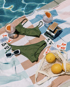 Ein freches Bikini-Top von Main Design in Olivgrün, eine weiße Sonnenbrille, Spielkarten und ein Grapefruit-Getränk liegen auf einem gestreiften Handtuch am Pool, daneben ein Netzbeutel mit Grapefruits und eine alte Sofortbildkamera.