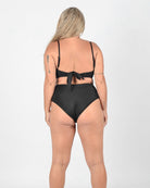 A person with long, blonde hair is standing barefoot facing away from the camera, modeling a black high-waisted bikini. The Adore Triangel Bikini Oberteil by Main Design with verstellbare Schulterträger has a knot tied at the back. They have several tattoos on their arms and legs, and the background is plain white.
