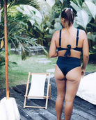 Eine Frau, die die schwarze Alice Bikini Hose von Main Design trägt, steht im Freien mit dem Rücken zur Kamera, neben einem weißen Liegestuhl und üppig grünen tropischen Pflanzen - ein stilvoller und nachhaltig inspirierter Look.