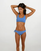 Eine Frau trägt das Charm Surf Bikini-Oberteil von Main Design in Blau und posiert vor einem weißen Hintergrund mit den Händen im Haar und betont UPF 50+ Sonnenschutz für optimalen Schutz beim Wassersport.