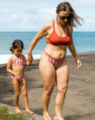 Eine Frau in einem roten Main Design Charm Surf Bikini Oberteil und einer gestreiften, chlorresistenten Hose hält die Hand eines jungen Mädchens in einem passenden UPF 50+ Badeanzug, während sie barfuß am Strand am Meer laufen.