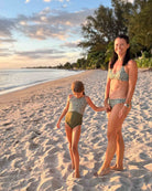 Eine Frau und ein junges Mädchen tragen passende Drift Surf Bikini Oberteile von Main Design, hergestellt aus nachhaltigen Materialien, während sie Hand in Hand an einem Sandstrand bei Sonnenuntergang mit Bäumen und dem Meer im Hintergrund stehen.  all-groups