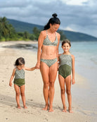Eine Frau und zwei junge Mädchen tragen passende Main Design Luxe Bikini-Hosen aus recycelten Materialien. Sie stehen barfuß an einem Sandstrand mit dem Meer und Palmen im Hintergrund, halten sich an den Händen und lächeln.  all-groups