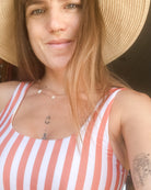 Eine Frau lächelt in die Kamera und trägt das Hale Bikini Oberteil von Main Design - ein rot-weiß gestreiftes, tragbares Badeanzug-Oberteil mit Rundhalsausschnitt. Dazu trägt sie einen Strohhut, eine Halskette, ein Septum-Piercing und zeigt ihre Tattoos.