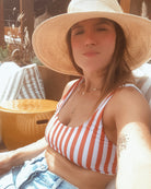 Eine Frau im Main Design Hale Bikini Oberteil mit rot-weißen Streifen und Jeansshorts entspannt sich an einem sonnigen Tag im Freien, sitzt auf einem Terrassenstuhl mit gestreiften Kissen, trägt einen breitkrempigen Hut und lächelt in die Kamera.