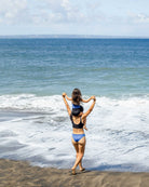 Eine Frau in einem blauen Main Design Hale Bikini Oberteil trägt ein Kind auf den Schultern, während sie am Sandstrand entlang spaziert, mit Blick auf die Wellen des Ozeans unter einem teilweise bewölkten Himmel.