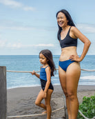 Eine Frau mit dem Hale Bikini Oberteil von Main Design und ein junges Mädchen in Badeanzügen stehen an einem Sandstrand. Die Frau trägt lächelnd das nachhaltig hergestellte Oberteil, während das Mädchen unter einem teilweise bewölkten Himmel einen Seilzaun hält.