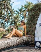 Eine Frau im Hale Bikini Oberteil von Main Design mit UPF 50+ Sonnenschutz sitzt lächelnd auf einer umgefallenen Palme an einem tropischen Strand, umgeben von üppigem Grün und einem aufrecht stehenden Surfbrett im Hintergrund.