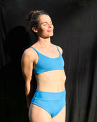 Eine Frau trägt das Sassy Bikini Oberteil reversibel von Main Design, hergestellt aus nachhaltigen Materialien. Sie steht vor einem schwarzen Hintergrund mit geschlossenen Augen, lächelt sanft und blickt ins Sonnenlicht.