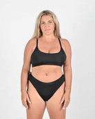 Eine Frau mit langen blonden Haaren steht selbstbewusst vor einem schlichten weißen Hintergrund und trägt das Sassy Bikini-Top von Main Design. Dieses schicke Stück besteht aus recycelten Materialien und hat einen nahtlosen Schnitt für maximalen Komfort und Stil.