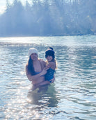 Eine Frau mit weißer Mütze und Badeanzug steht in einem Fluss und hält ein lächelndes Kind, das den Lena Mädchen Badeanzug von Main Design (UPF 50+) trägt. Das Sonnenlicht glitzert auf dem Wasser, umgeben von Bäumen und leichtem Morgennebel.