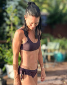 Eine Frau mit nassen dunklen Haaren trägt das Sassy Bikini-Top von Main Design und steht im Freien im Sonnenlicht und schaut mit einem leichten Lächeln nach unten. Der Hintergrund ist mit Grünzeug und Terrassenmöbeln unscharf.