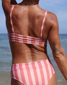 Eine Person steht am Strand mit dem Rücken zur Kamera und trägt das Main Design Sassy Bikini-Top in rosa und weißen Streifen. Ihre gebräunte Haut und ihr Tattoo sind sichtbar, mit dem Meer und dem Himmel als schöne Kulisse.