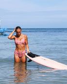 Eine Frau, die das Main Design Sassy Bikini-Top trägt, steht im flachen Wasser des Ozeans, hält ein rosa Surfbrett in der Hand und berührt ihr Haar, während das Meer ruhig ist und der blaue Himmel im Hintergrund zu sehen ist.