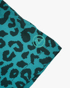 Nahaufnahme des blaugrünen Stoffes mit dunklem Leopardenmuster und 4-Wege-Stretch. Ein kleines, rundes Logo mit einem umrandeten Dreieck befindet sich am Rand, ideal für die Lean Boardshorts für Jungen von Main Design mit UPF 50+ Sonnenschutz.