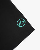 Nahaufnahme des schwarzen 4-Wege-Stretch-Stoffes der Lean Boardshorts für Jungen von Main Design mit einem grün gestickten Logo, das einem stilisierten Dreieck in einem Kreis ähnelt. Sauber vernähte Kanten sorgen für Strapazierfähigkeit und Stil.