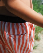 Ein Kind trägt Main Design Lean Boardshorts für Jungen in rot-weiß gestreift mit einem schwarzen Bund. Die schnell trocknenden Shorts bieten UPF 50+ und sind im Freien abgebildet, mit grünen Pflanzen, die im Hintergrund sanft verschwimmen.